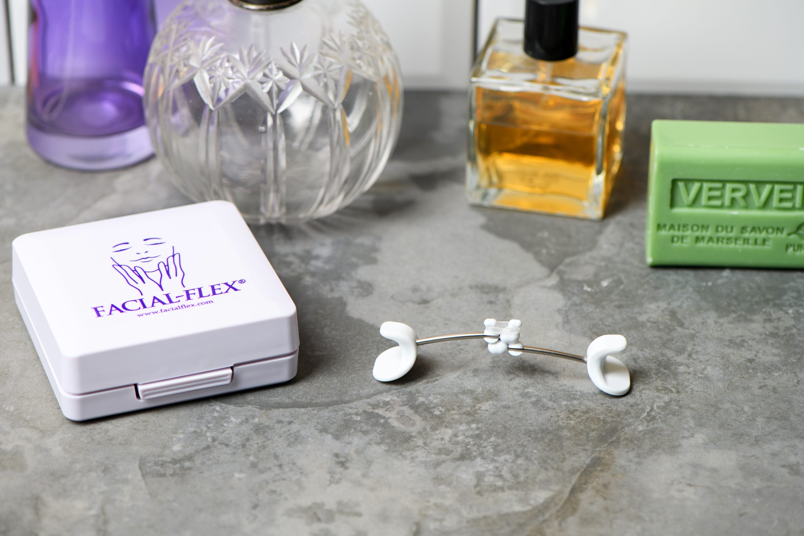 A Facial Flex sitting on a dressing table beside its carry box alongside several cut glass perfume bottles and a bar of facial soap.